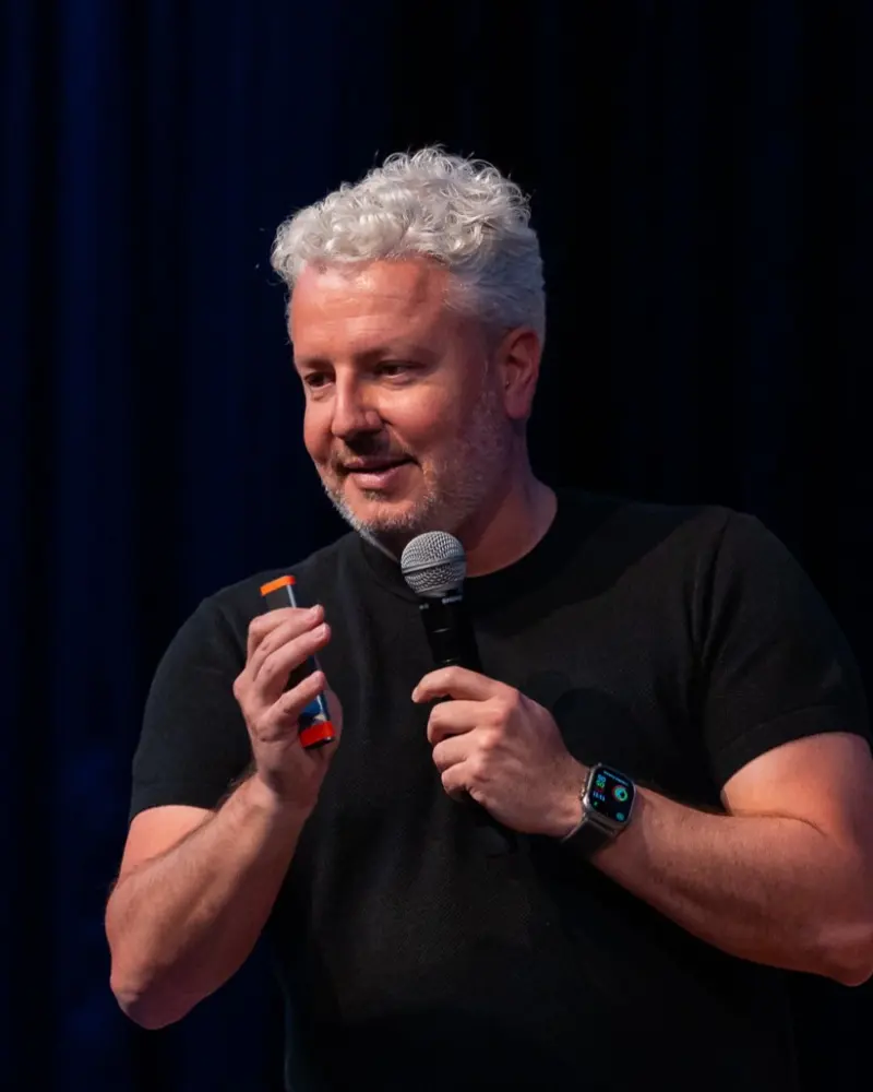 Ricardo Wagner in a black T-shirt smiling with a microphone on stage.